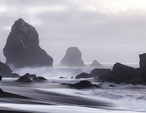 Dark sea stacks and long-exposure surf on a black sand beach under gray sky